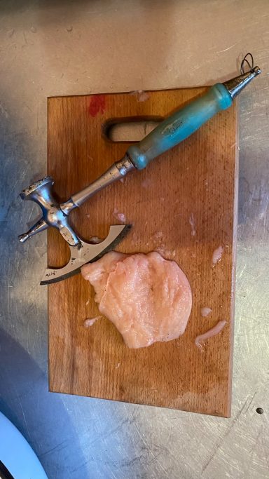A wooden cutting board with a metal meat tenderizer and flattened meat.